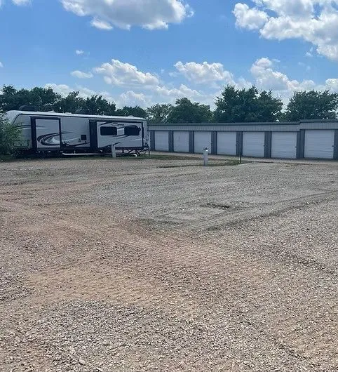 A large RV parked on a gravel lot next to a row of white storage units under a blue, cloudy sky.